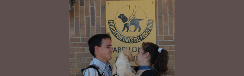 Two people hold a puppy in front of a sign for guide dog training school in Spain