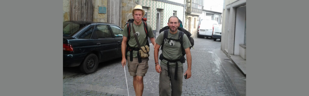 Nick walks with cane and his friend through narrow street in Spain.