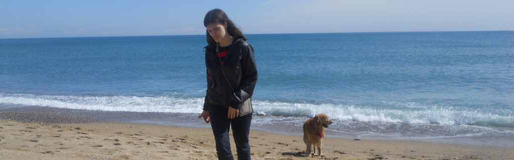 A young American woman walks along a beach with a dog.