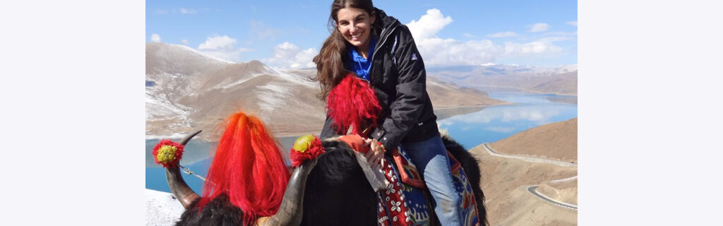 A young woman sits atop a wooly yak with a lake and mountains in the background.