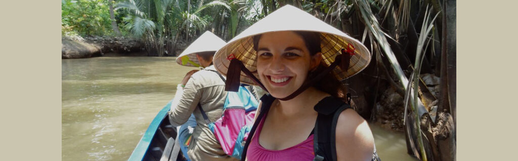 Emily block wears a Vietnamese-style hat while riding in a boat on a river.