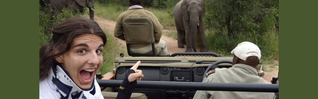 A young woman wearing a neck support excitedly points at an elephant from a safari car.