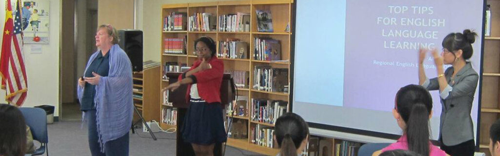 A middle-aged woman addresses an audience while two younger women stand behind her to interpret in sign language. A screen behind them reads "Top Tips for English Language Learning."