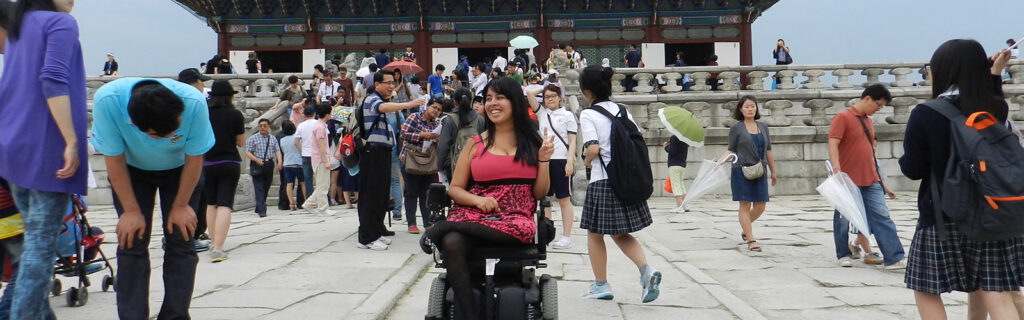 A young American woman in a power wheelchair in a busy square in an Asian city.