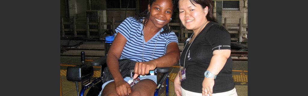 Two exchange participants of diverse backgrounds smile in the camera: A power wheelchair user and a woman of short stature