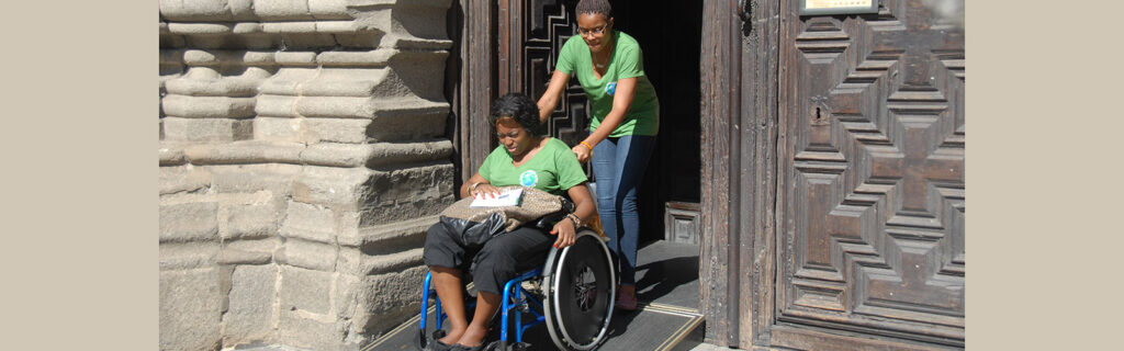 Woman in a manual wheelchair gets assistance down a ramp outside a carved wooden door