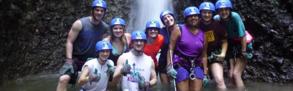 Study Abroad students including one who is blind pose with their hard hats on at the bottom of a waterfall in Costa Rica they ziplined by.