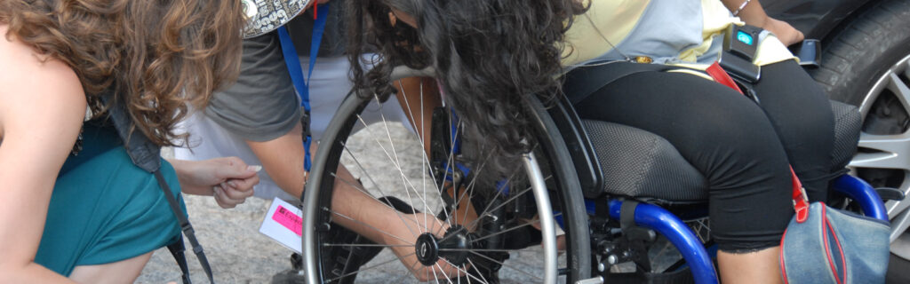 Three people, including the wheelchair user, lean over to check out the axel of her manual wheelchair