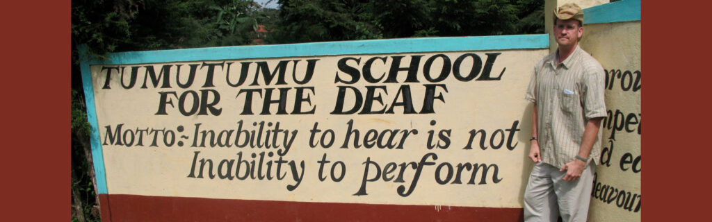 A man stands next to a hand-painted sign that reads "Tumutumu School for the Deaf, Motto: Inability to hear is not inability to perform"