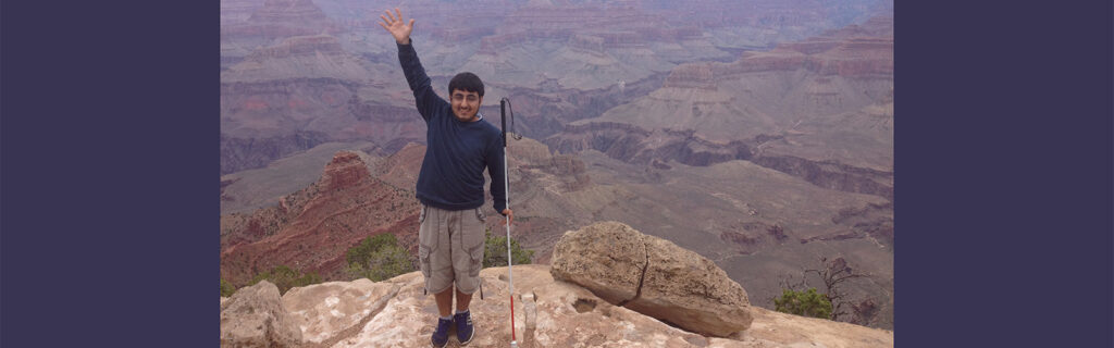 International student who is blind standing on top of the Grand Canyon smiling with white cane in his hand.