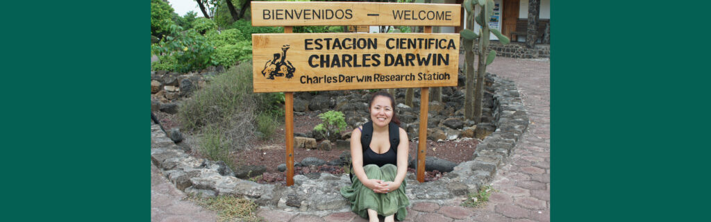 Mayuko sitting in front of the Charles Darwin Research Station in Ecuador and sign that reads "Estacion Cientifica Charles Darwin".