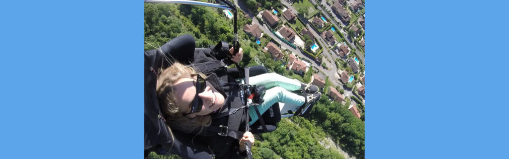 Floriane paragliding over many trees, houses, and pools below her.