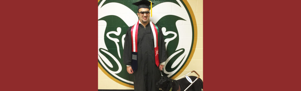 International student, who is blind, stands wearing his graduation cap and gown, with his guide dog next to him.