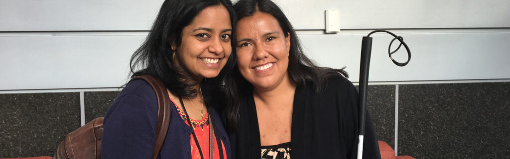 Close up of two women leaning heads together and smiling. Woman on right holds a white cane.