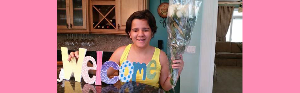 Seated at a kitchen table, exchange student Pinar holds a bouquet of flowers in one hand while supporting a sign that reads "Welcome" with her other. She is smiling and wearing a bright yellow tank top.