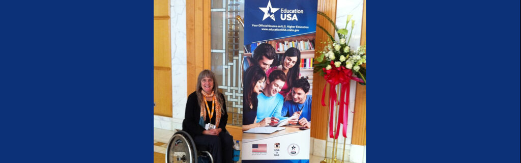 MIUSA CEO and wheelchair rider, Susan Sygall, smiles next to EducationUSA banner.