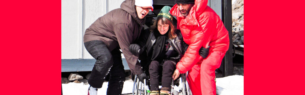 Two men on each side lift the frame of Susan's wheelchair up a short step