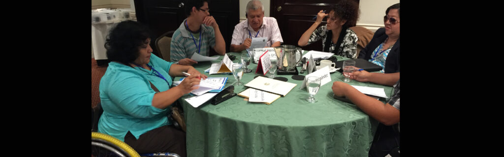 Five people are seated around a table; the woman on the left is a wheelchair user; next to her are two men discussing a document; to their right are two women--one has sunglasses on