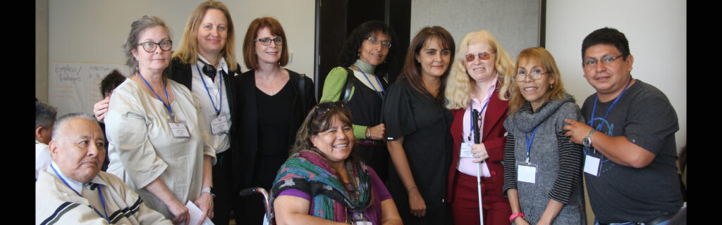 Seven women (one with a cane) and one man stand and face the camera; a wheelchair user is seated at center--she is smiling