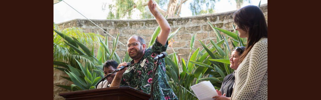 Jacob speaking behind podium with one arm in the air and others on stage smiling , looking on.