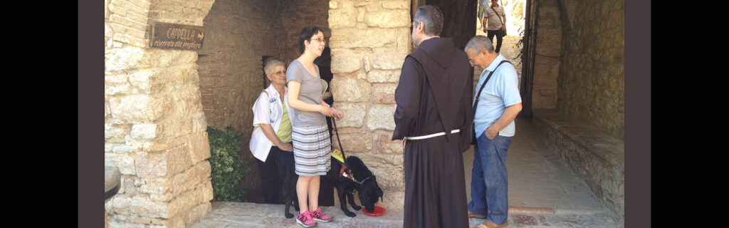 Jameyanne speaking with a priest in front of the arch of an old cathedral with her guide dog by her side.