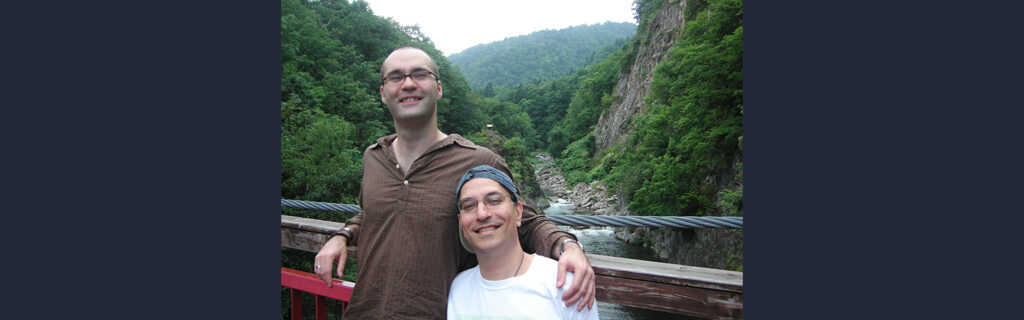 Kenny standing on bridge with a friend overlooking a river and rolling green hills in the background.