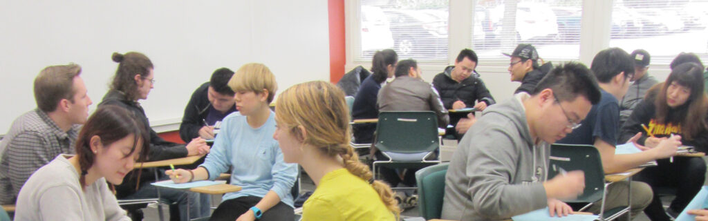 Room full of students talking and sitting at their desks in small groups with blue books on their desk and pens in hand.