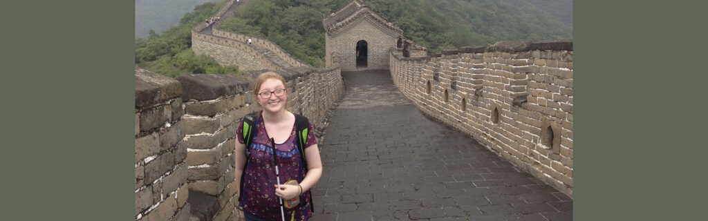 A young white woman holds a white cane. Behind her is the Great Wall of China