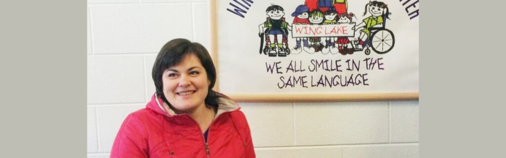 A smiling woman looks off in the distance. Behind her is a sign that says "We all smile in the same language" with images of children with and without disabilities.