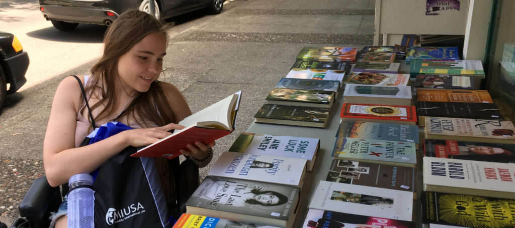 Student using a powerchair browsing books for sale on a table outside a shop