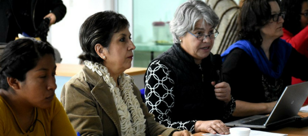 Members of RightsNow and the Mexican organization MADIJAL sit at a conference table together during a planning meeting. Susei Grimes speaks into a microphone and is surrounded by at least five more women.