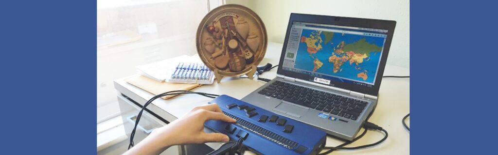 A laptop displays a world map. A hand reads the braille display attached to the laptop at a desk.