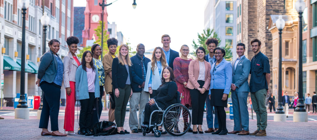 A group of a dozen young people, including men and women of color, a student seated in a wheelchair, and a blind person with a service dog at her feet. Together, they gather in an outdoor urban setting.