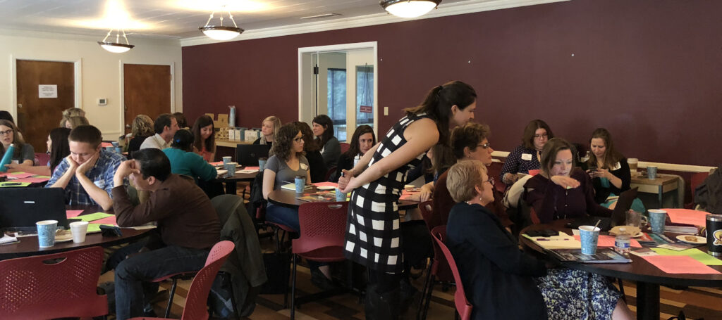 In a room full of people seated around round tables, one standing woman leans over a group to join their discussion