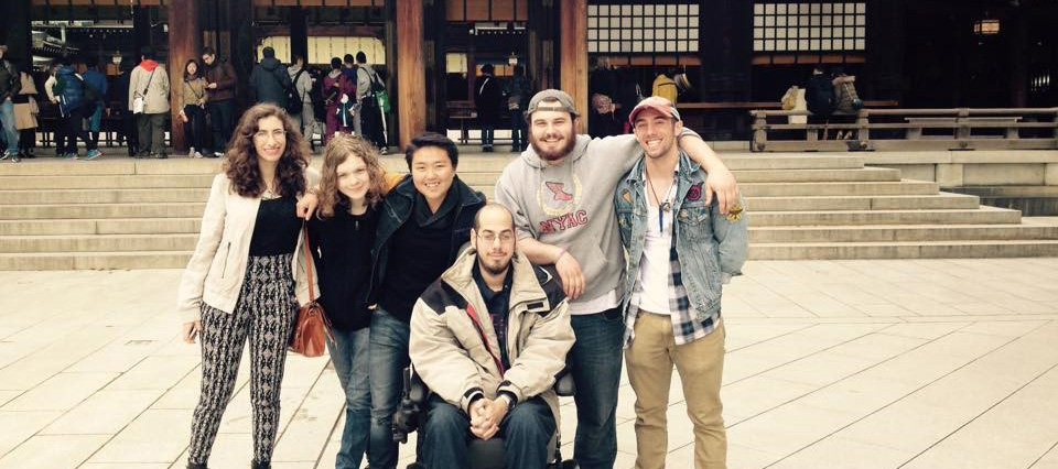 A group of men and women, including a man seated in a power wheelchair, put arms around shoulders in front of a Japanese shrine