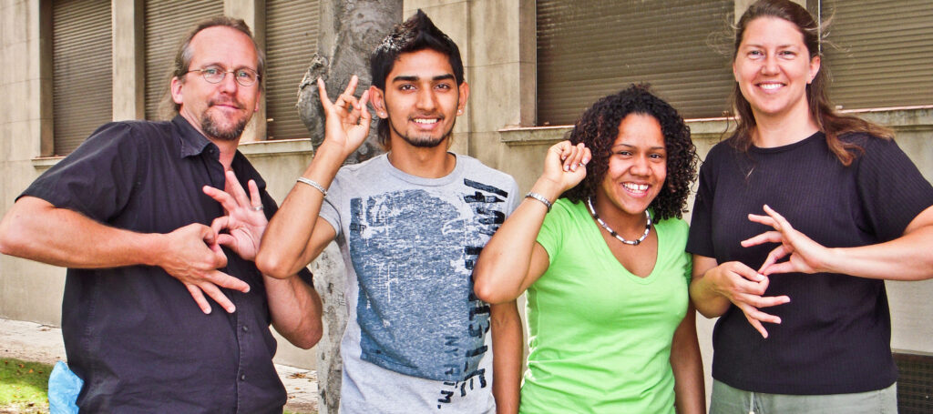 Four people standing outdoors smile at camera while displaying ASL signs on their hands. An older white man and woman make sign for "interpreter" while two younger people of color show their name signs.