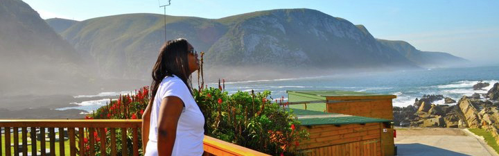 A young woman wearing sunglasses stands on a wooden deck that overlooks the ocean. She faces sideways. Mountains are visible in the background.