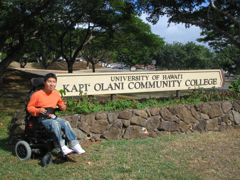 Student, who uses a power wheelchair, poses next to "University of Hawaii Kapi'Olania Community College" sign outside.