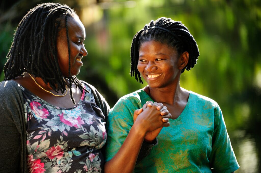 Two young black women face each other, smiling, as they clasp hands and stroll through a forested green landscape