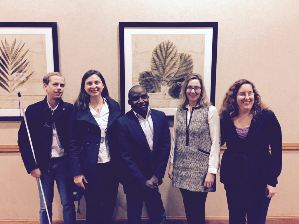 Five individuals stand next to each other in a hallway: a white man holding a white cane; a young white-appearing woman, a black man of short stature; a white woman, and a white woman who is low vision