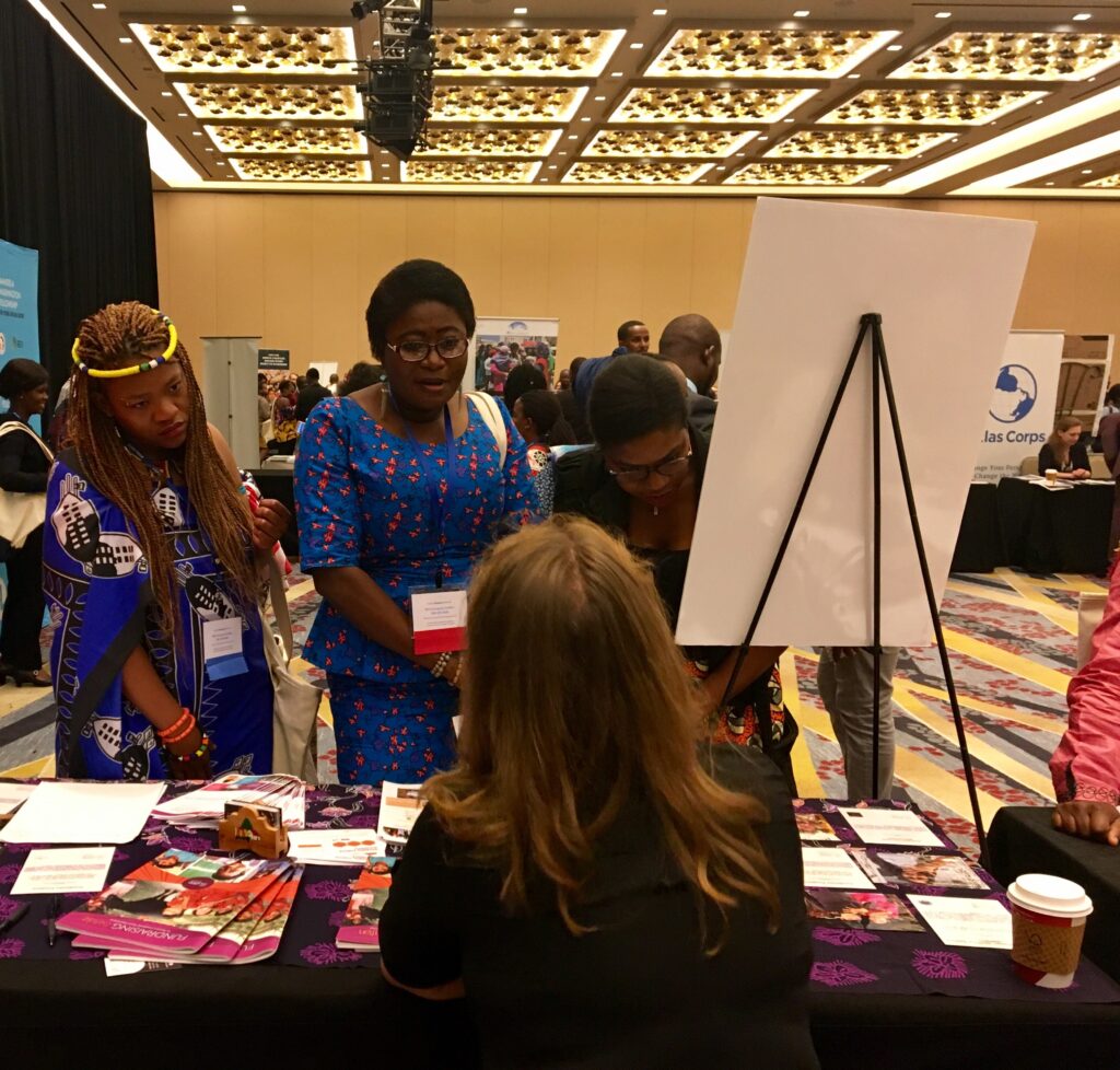 Three women wearing traditional African clothes stand at an info table in a large expo hall with another woman sitting behind table.