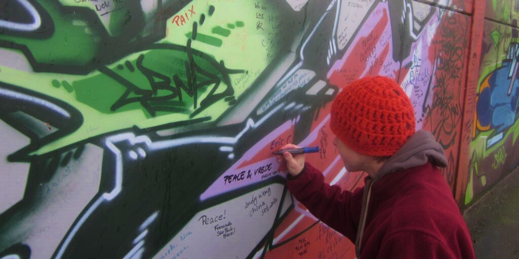 A young man, viewed from behind, uses a purple marker to write "Eric C" on a colorful wall that has been airbrushed with green, black and pink patterns. He wears a knit cap and sweatshirt.