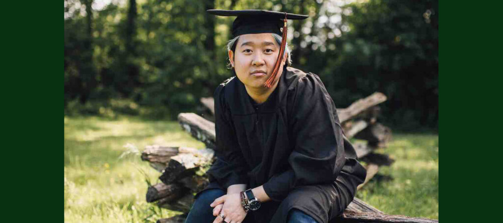 Close up of a young man of southeast Asian origin wearing graduation cap and gown and sitting in forested setting. He wears a neutral expression.