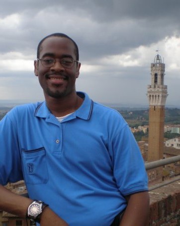 Joseph leans against a balcony railing in Italy, with tower in background