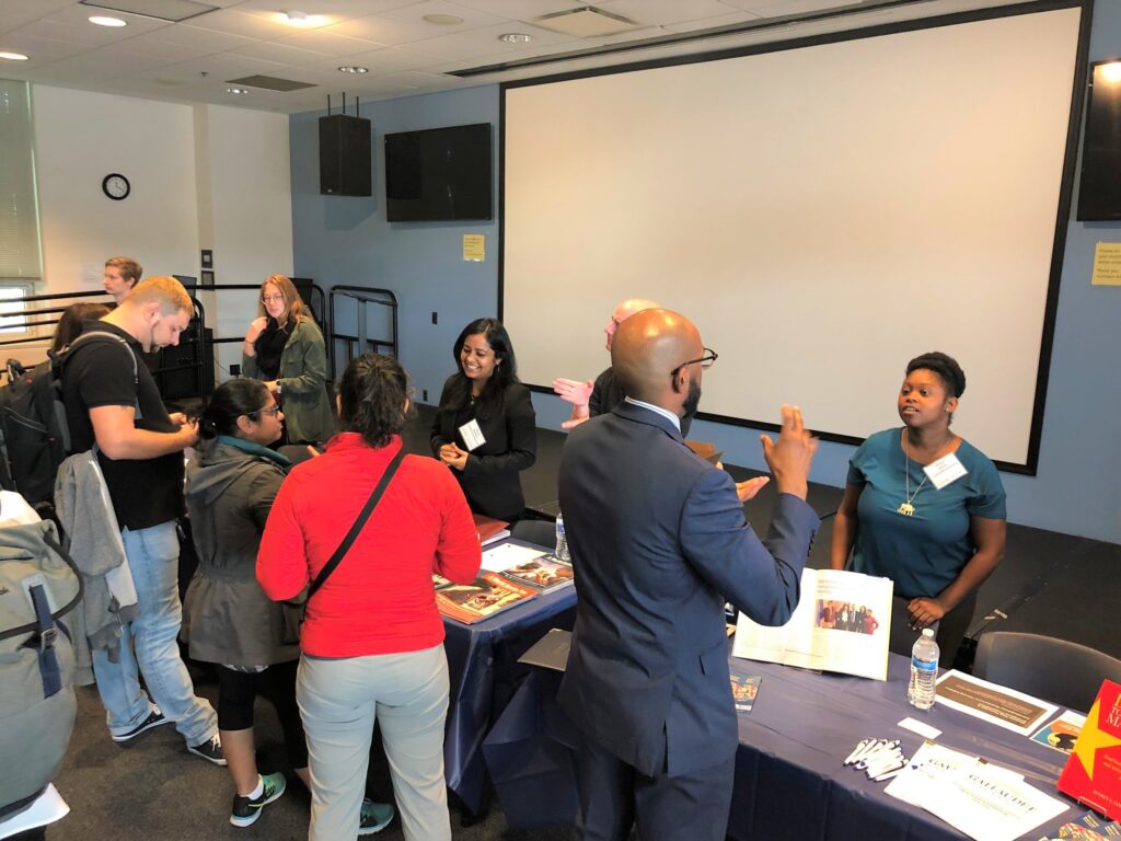 People mill around two exhibit tables and speak or sign questions with staff inside a large exhibit hall