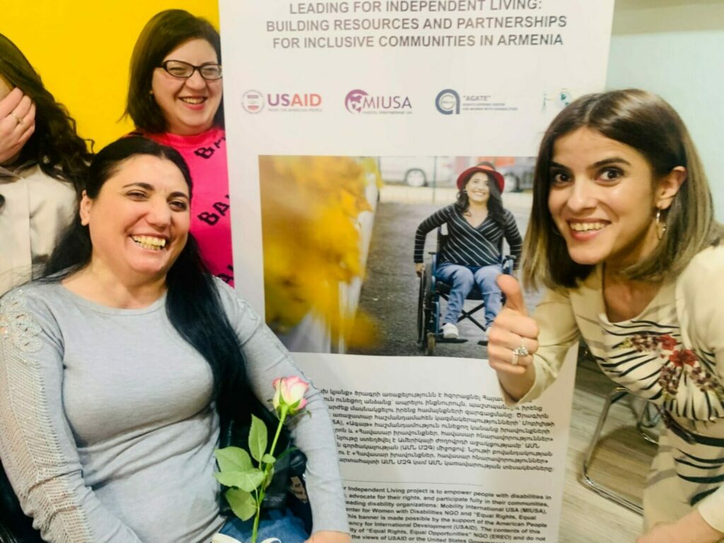 Two women are smiling at the camera. The woman on the left is a wheelchair rider and holds a rose, and the woman on the right is showing a thumbs up to the camera. Behind them is a project banner that says Leading for Independent Living: Building Resources and Partnerships for Inclusive Communities in Armenia.
