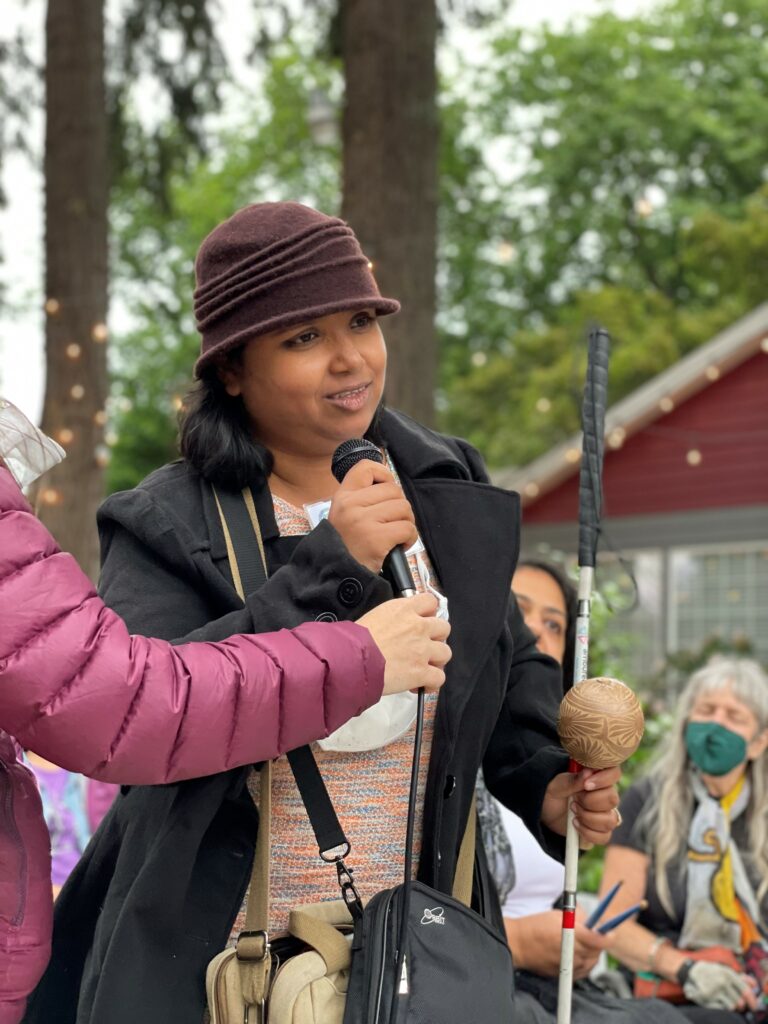 A blind woman holds a white cane and percussive instrument in one hand, and a microphone in the other as others listen.