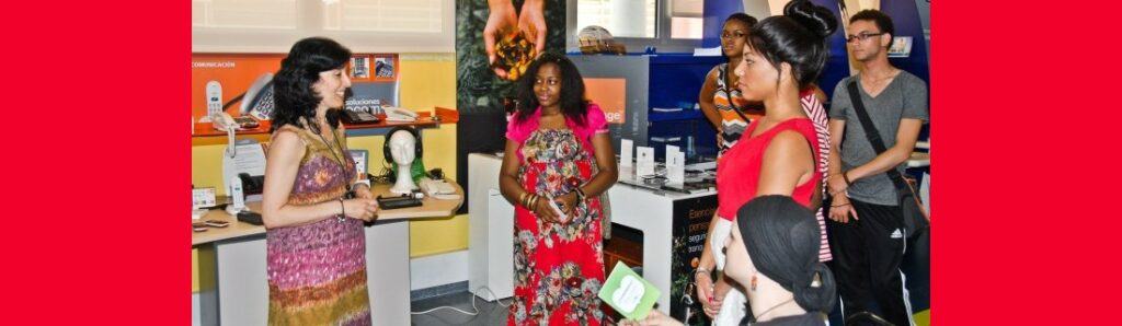 A small group of young people stand or sit watching a woman who is presenting to them inside an office.