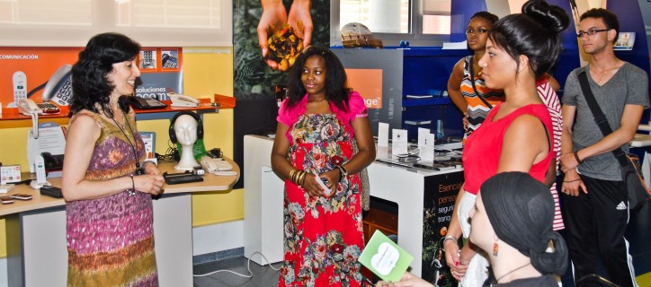 A small group of young people stand or sit watching a woman who is presenting to them inside an office.