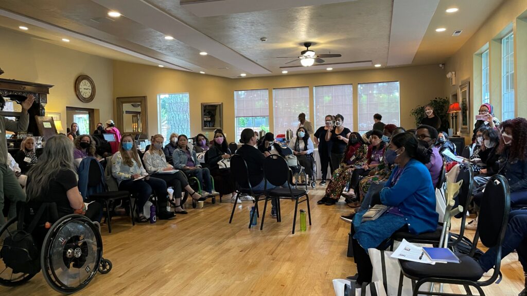 About 50-60 people gathered in a circle in an indoor conference room, seated in a semi circle. Susan Sygall is seen facing the crowd.